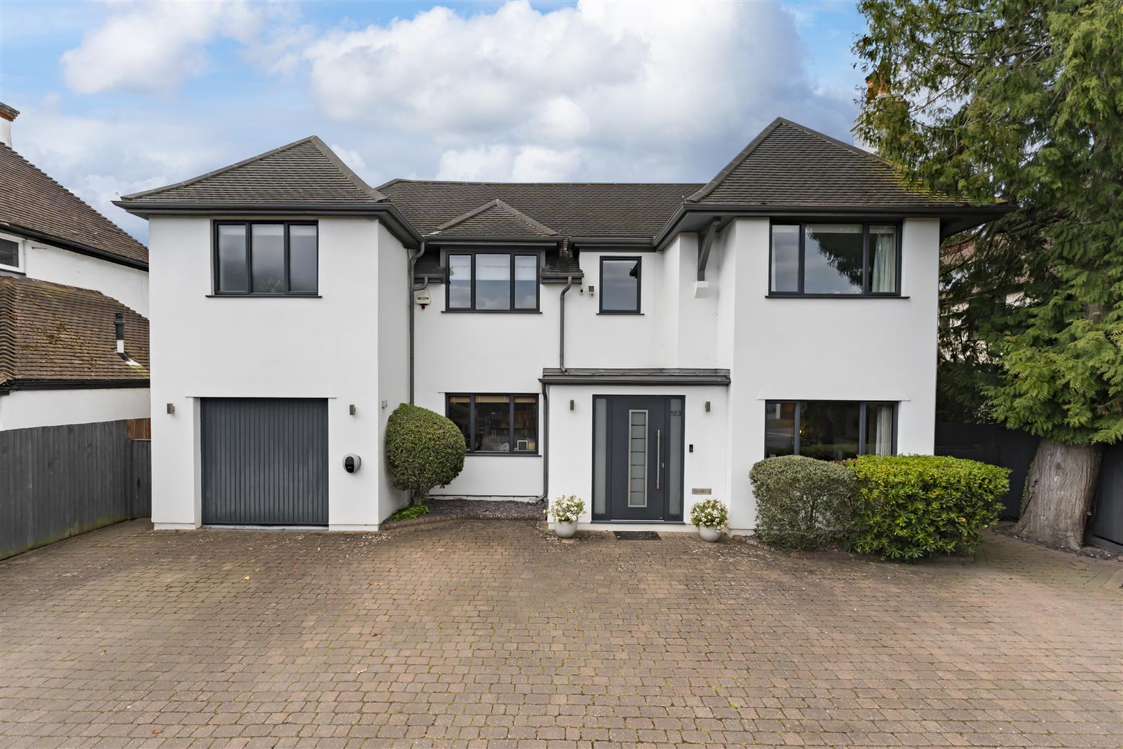 White two-story house with a grey roof and attached garage, front door centered between trimmed shrubs on a brick driveway.