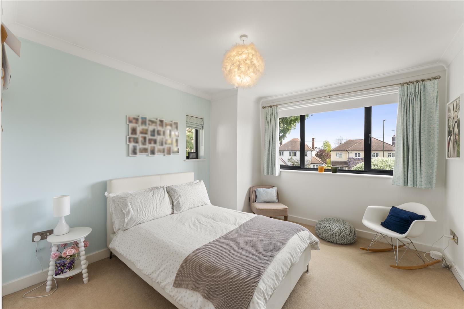 Bright bedroom with a light blue accent wall, white bed linens, and a large window offering a view of houses outside; cozy seating area nearby.