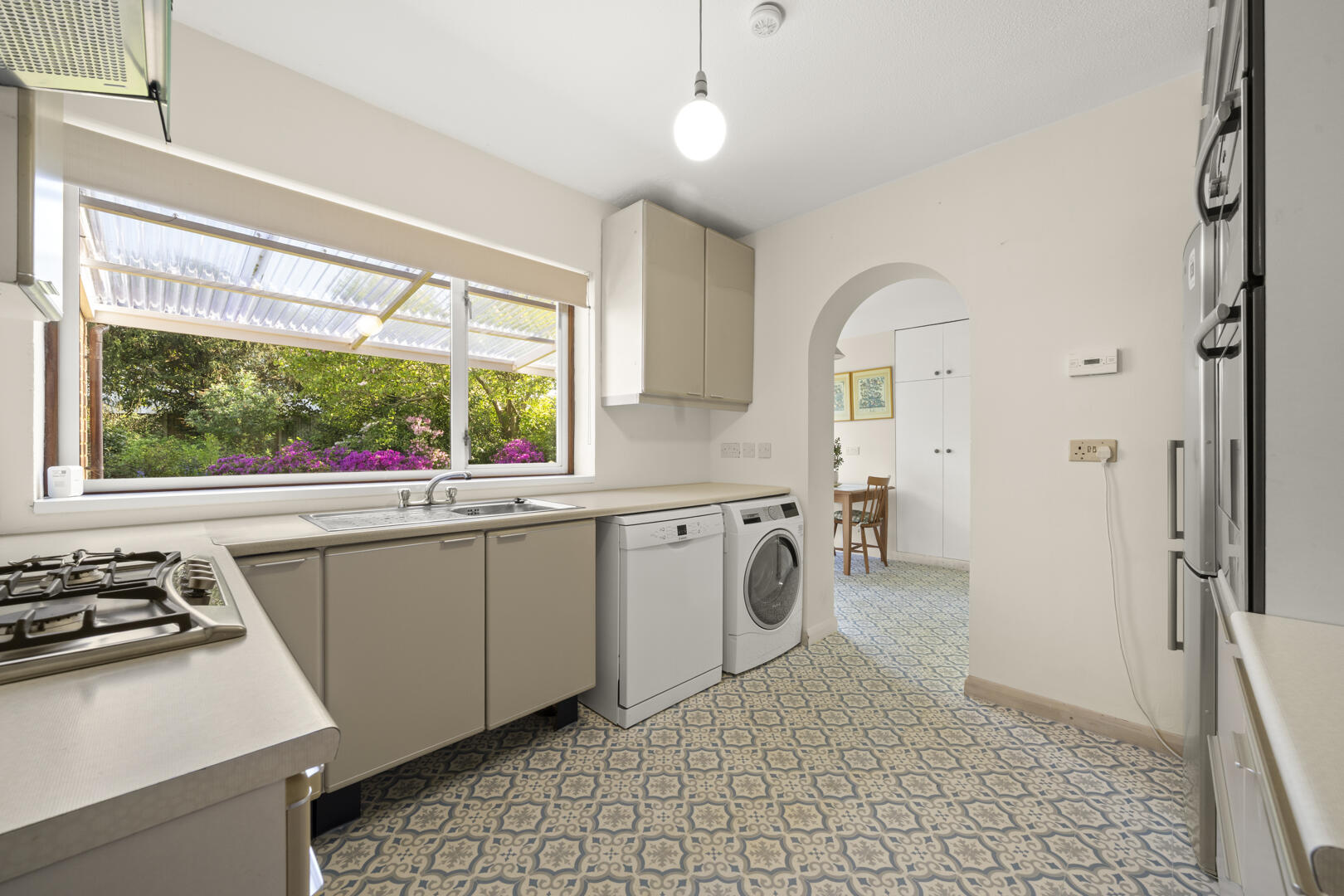 Bright kitchen with a large window overlooking a garden, beige cabinets, sink, stove, and a row of laundry appliances nearby under a hanging bulb.