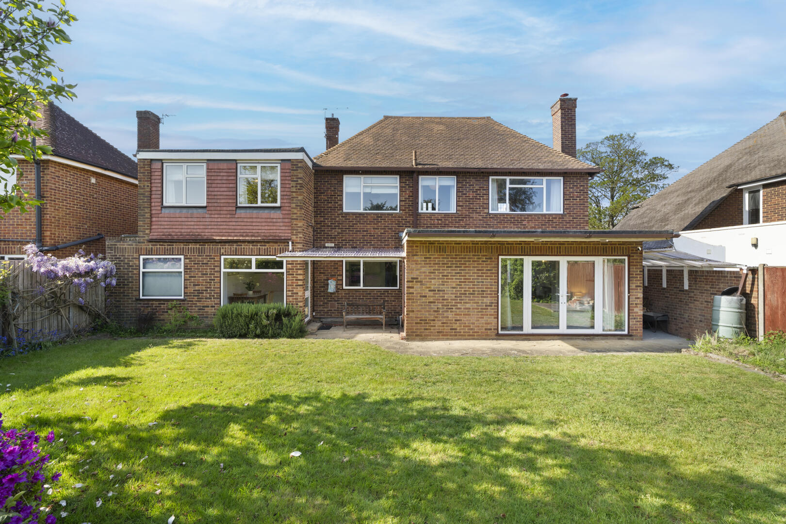Back view of a brick two-story house with a well-kept lawn, a paved patio, and sliding glass doors leading to an extension. A wooden fence lines the left yard with purple wisteria in bloom.