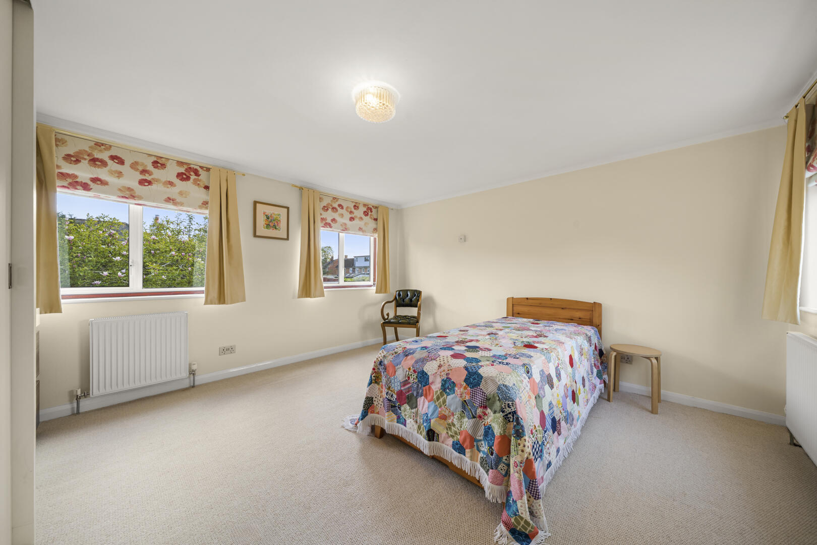 Bedroom with a single wooden bed and colorful patchwork quilt, beige walls, and cream carpet; three windows with floral blinds and a chair nearby.