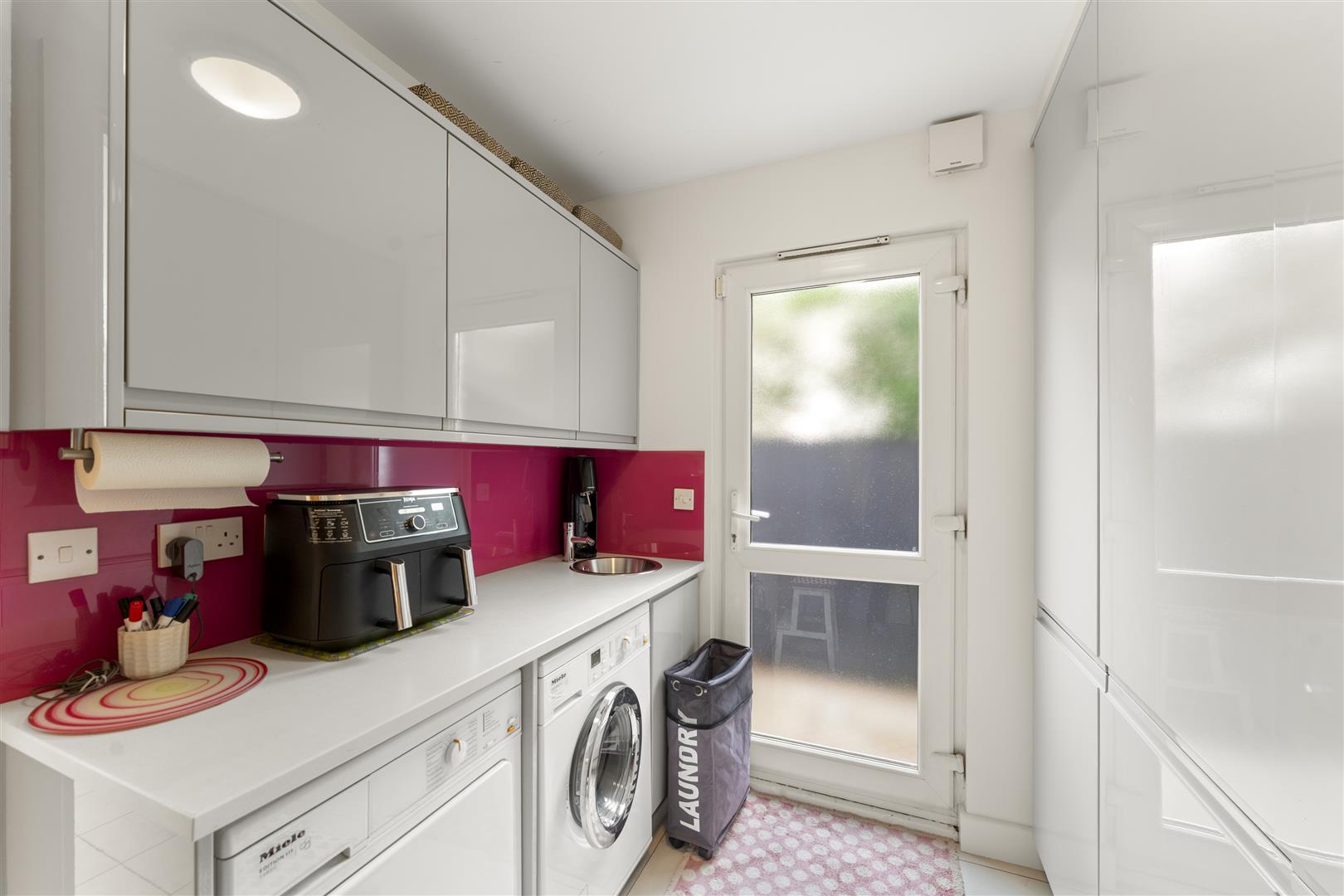Bright white laundry room with glossy cabinets, pink backsplash, washing machine and dryer under counter, and a sink to the right of the appliances.