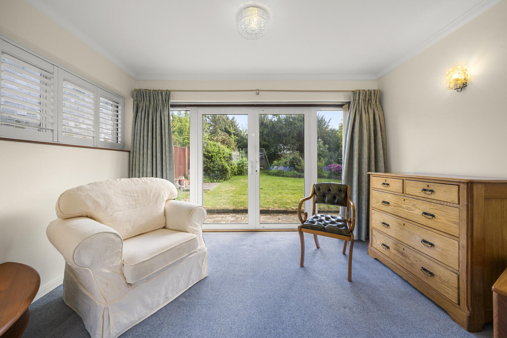 Living room with a cream upholstered armchair, wooden dresser, and large glass doors opening to a garden.