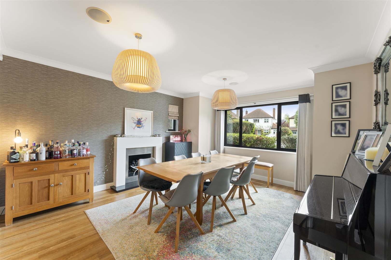 Contemporary dining room with a wooden table and gray chairs, a bar cabinet with bottles on the left, and a glossy piano on the right, glass sliding windows in the background.
