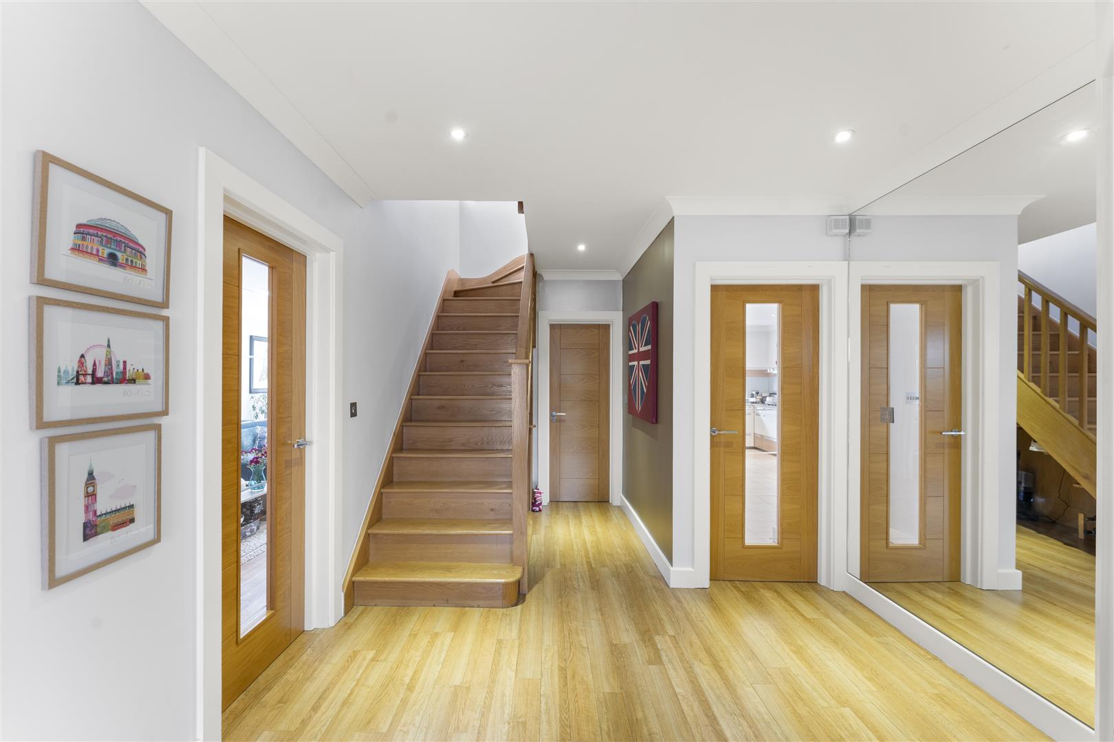 Bright hallway with wooden stairs to the left, light gray walls, and multiple wooden doors; framed pictures on the left wall and a Union Jack art piece on the right wall.