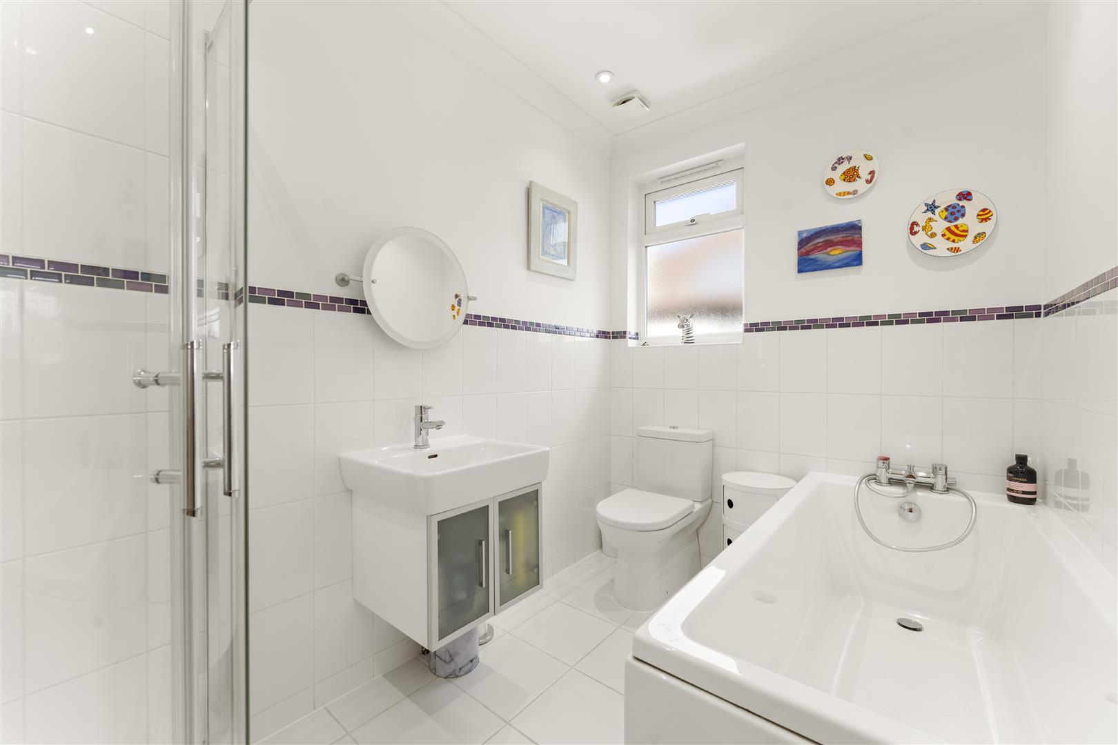Bright white bathroom with a glass‑front shower, pedestal sink, and bathtub along the right; purple mosaic tile line adds color.