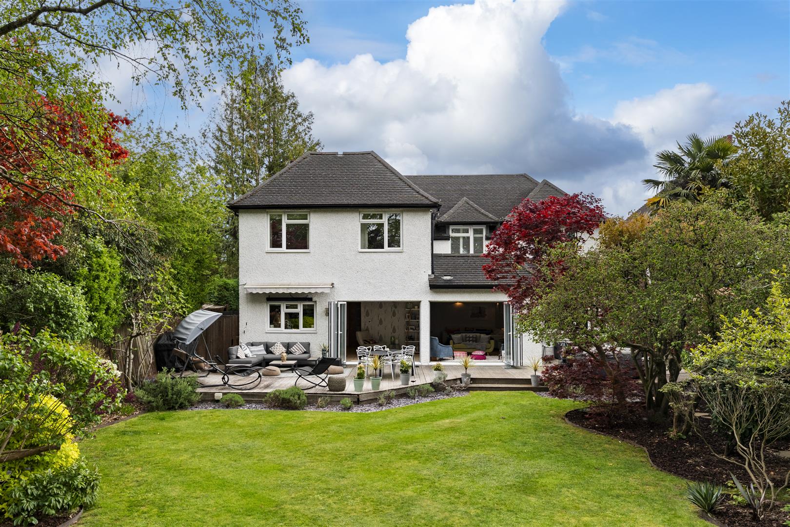 White two-story house with a tiled roof and a spacious patio area with outdoor seating and potted plants in a lush garden setting.
