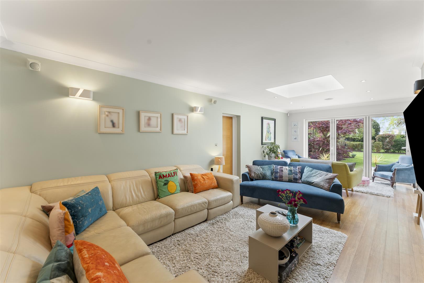Bright living room with pastel green walls, cream sectional sofa, blue sofa, and colorful throw pillows; large glass doors open to a green garden.
