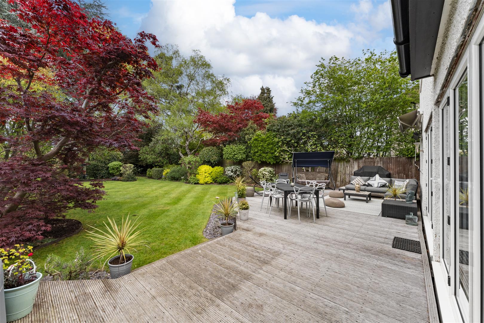 Backyard with a wooden deck, outdoor dining table, chairs, and a seating area amid green lawn and colorful trees.