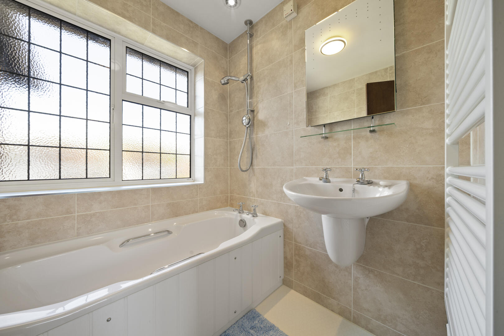 Bathroom with a white tub along the left, beige tiled walls, and a frosted grid window; pedestal sink nearby and a wall mirror above a glass shelf.