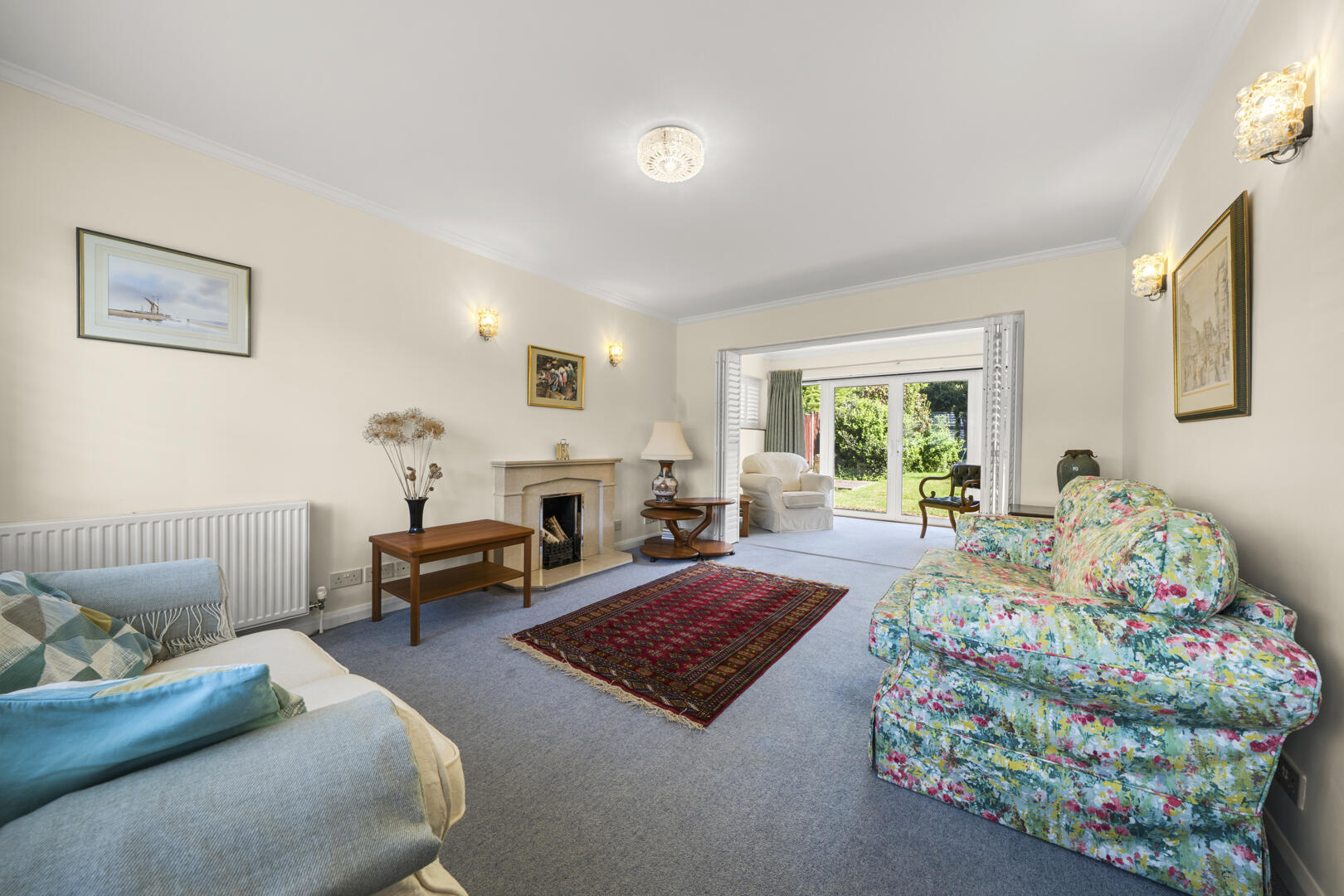 Bright living room with cream walls, a floral-patterned sofa, blue cushions, and a central red rug by a fireplace. Sliding doors open to a sunny garden.