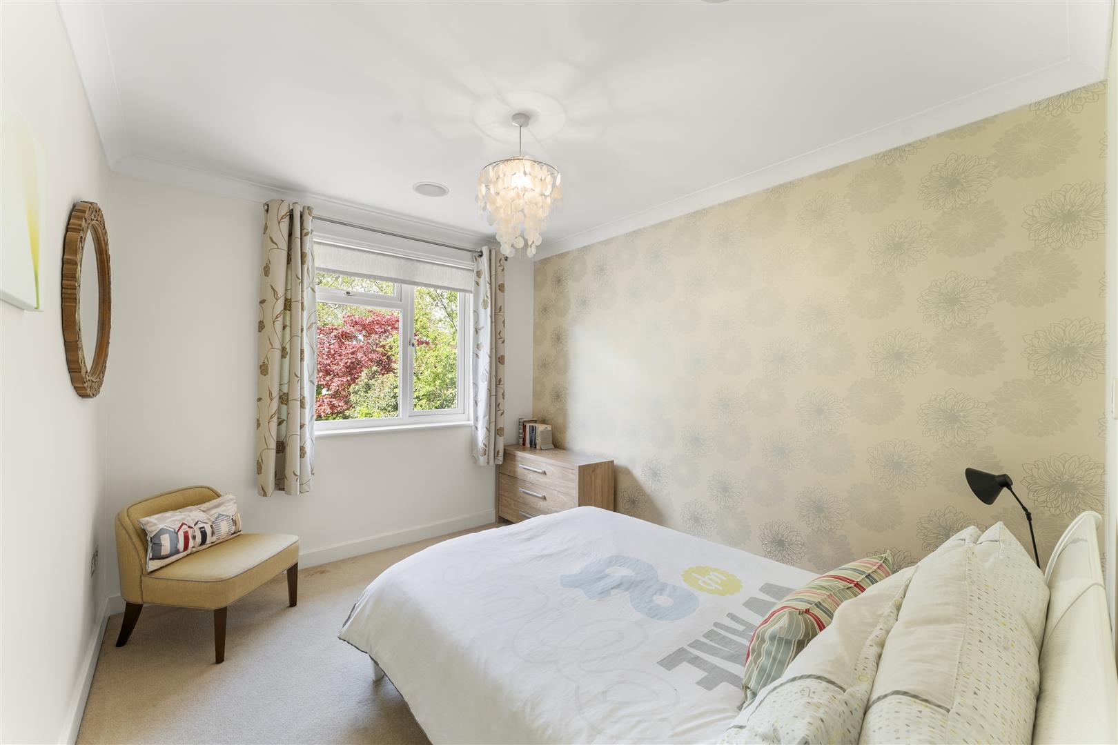 Bright bedroom with a window, floral curtains, bed in foreground, and a wooden dresser against a pale patterned wall.