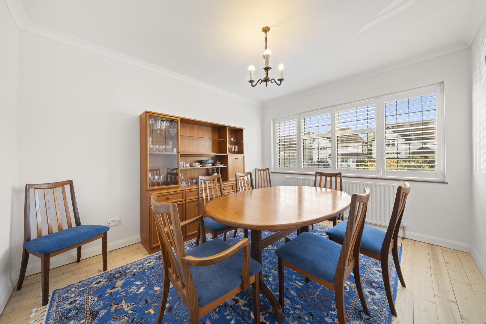 Bright dining room featuring a round wooden table surrounded by chairs with blue cushions, a wooden china cabinet, and a blue patterned rug underfoot; large windows with white blinds let in daylight; a chandelier hangs from the ceiling.