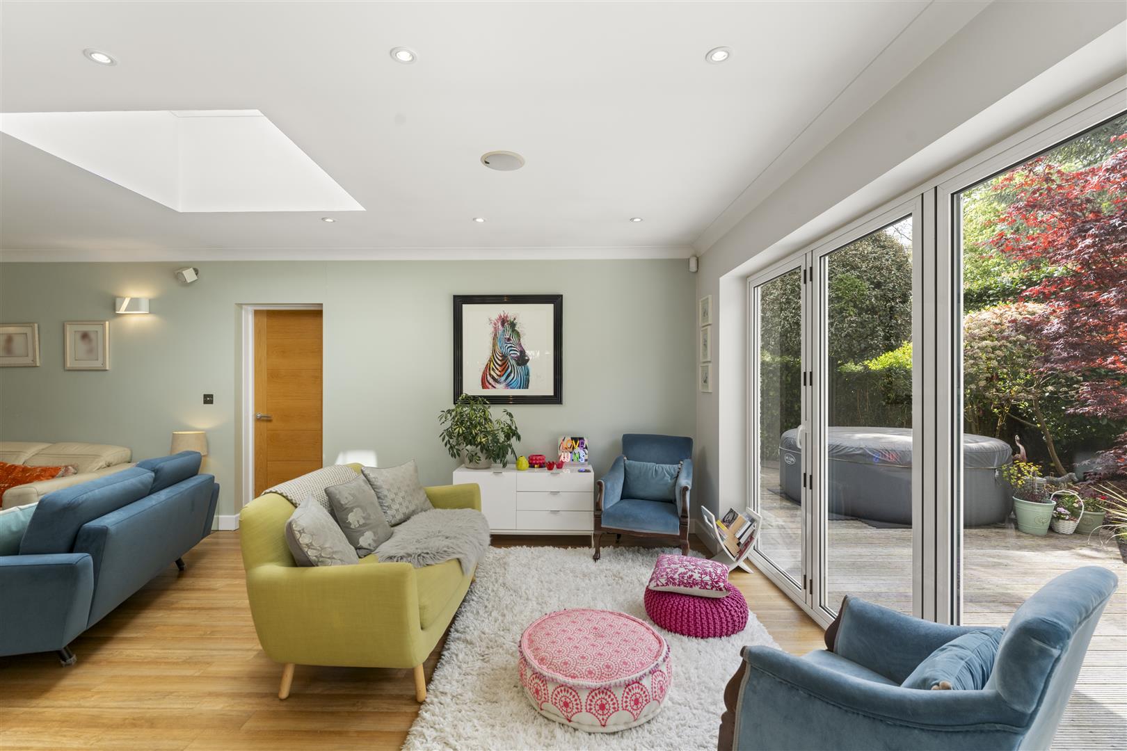 Bright living room with pastel furniture, white rug, and large sliding glass doors opening to a garden with a patio and hot tub outside.