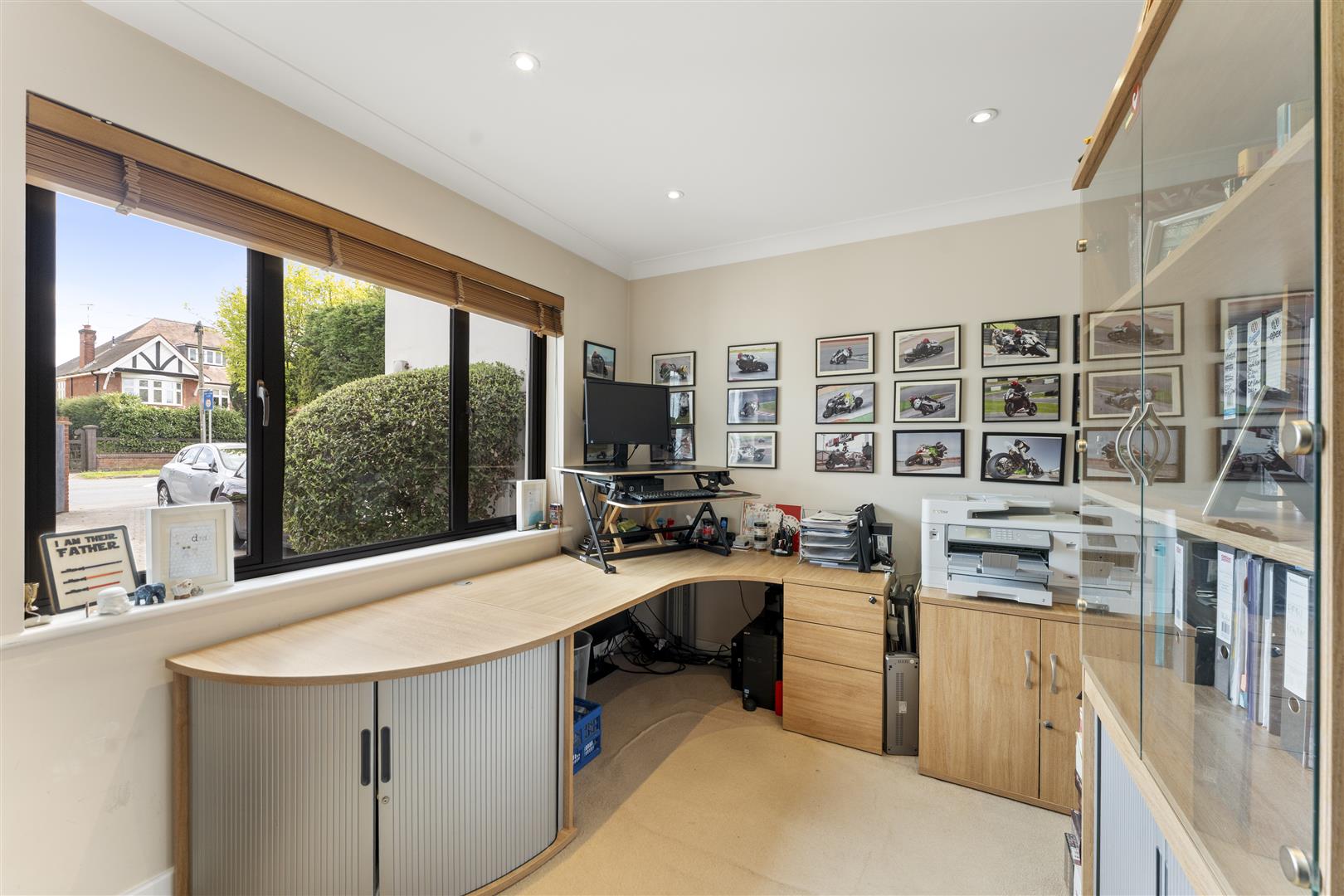 Home office with a curved wooden desk, monitor on stand, and a wall of framed motorcycle photos behind the desk.