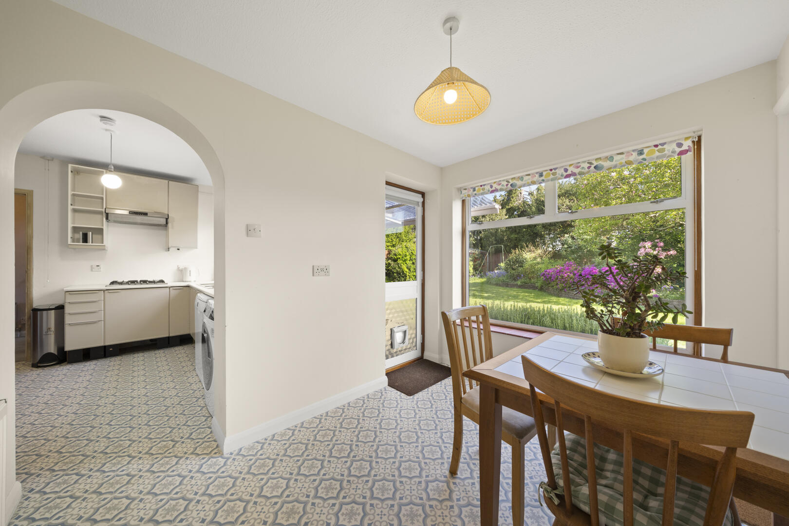 Bright dining area with a white-tiled table, wooden chairs, and a potted plant, overlooking a lush garden through a large window; arch to a small kitchen.