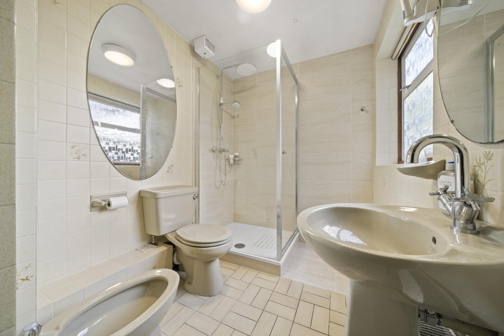 Bright beige-tiled bathroom with toilet, bidet, pedestal sink, and glass shower enclosure; oval mirror overhead and a window to the right.