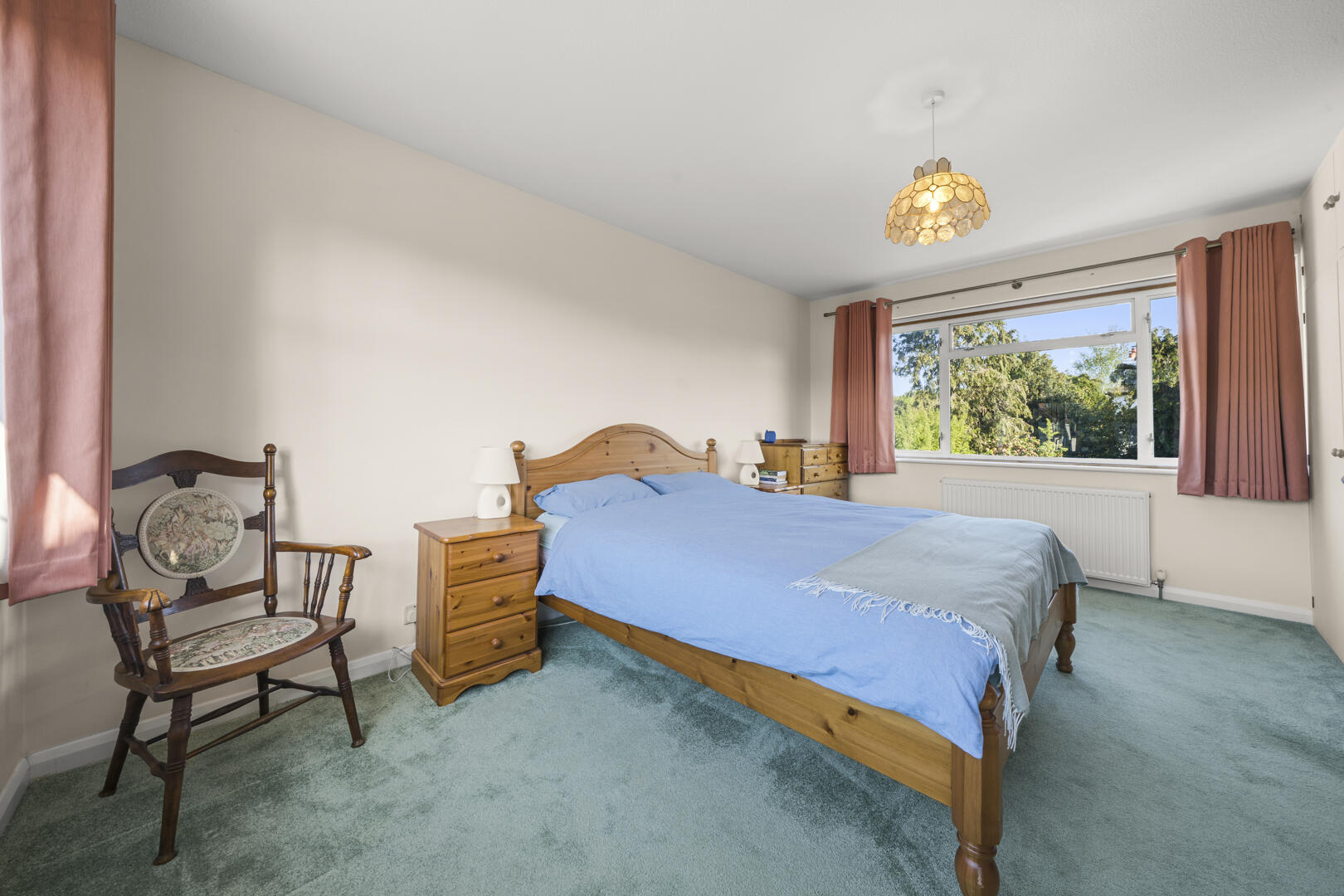 Cozy bedroom with a wooden bed, blue bedding, matching nightstands, and a dresser by a large window with pink curtains.