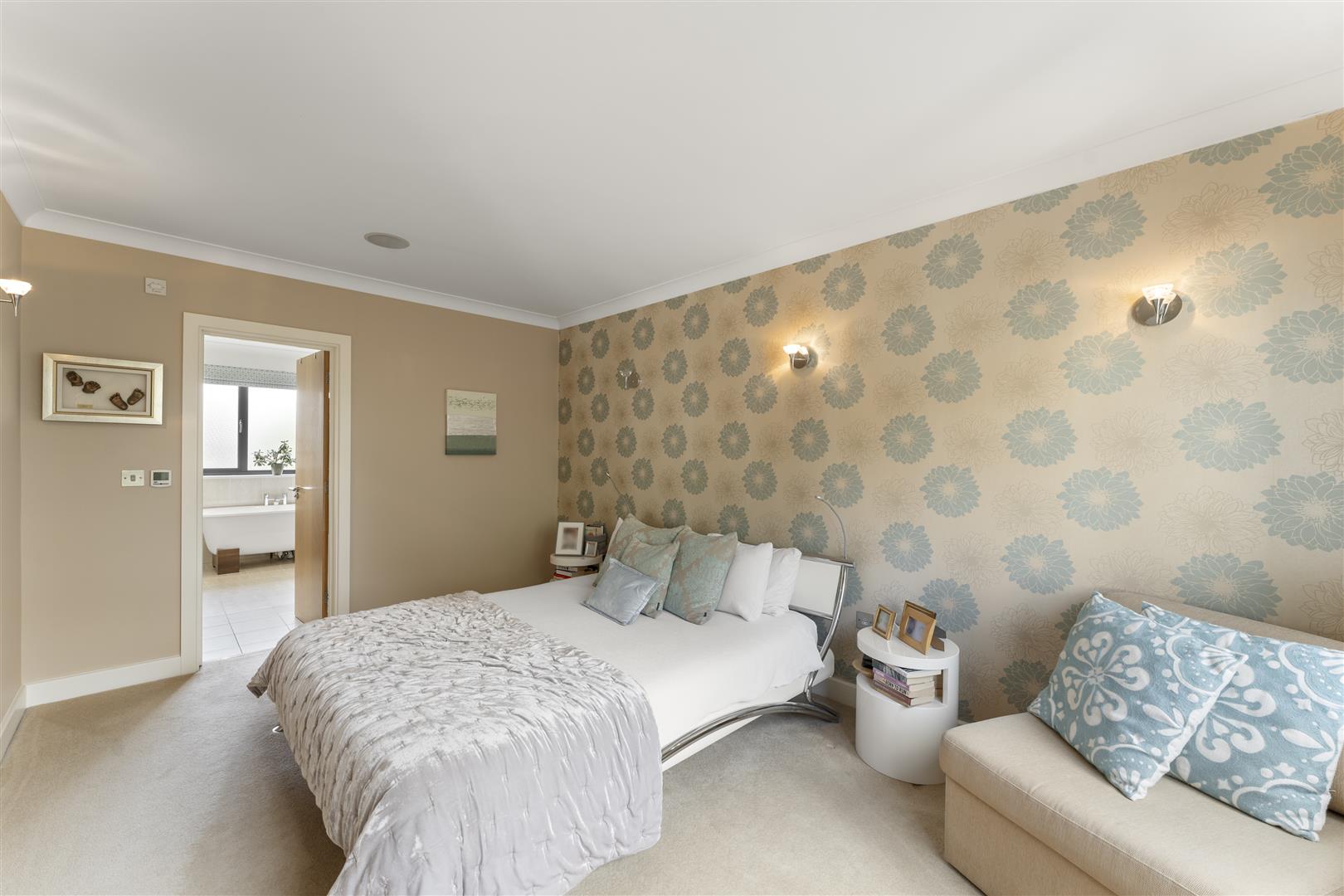 Bedroom with beige walls and a blue floral wallpaper accent wall, a neatly made bed with gray quilt and pastel cushions, and a cream sofa with blue patterned pillows; doorway to a bathroom visible on the left.