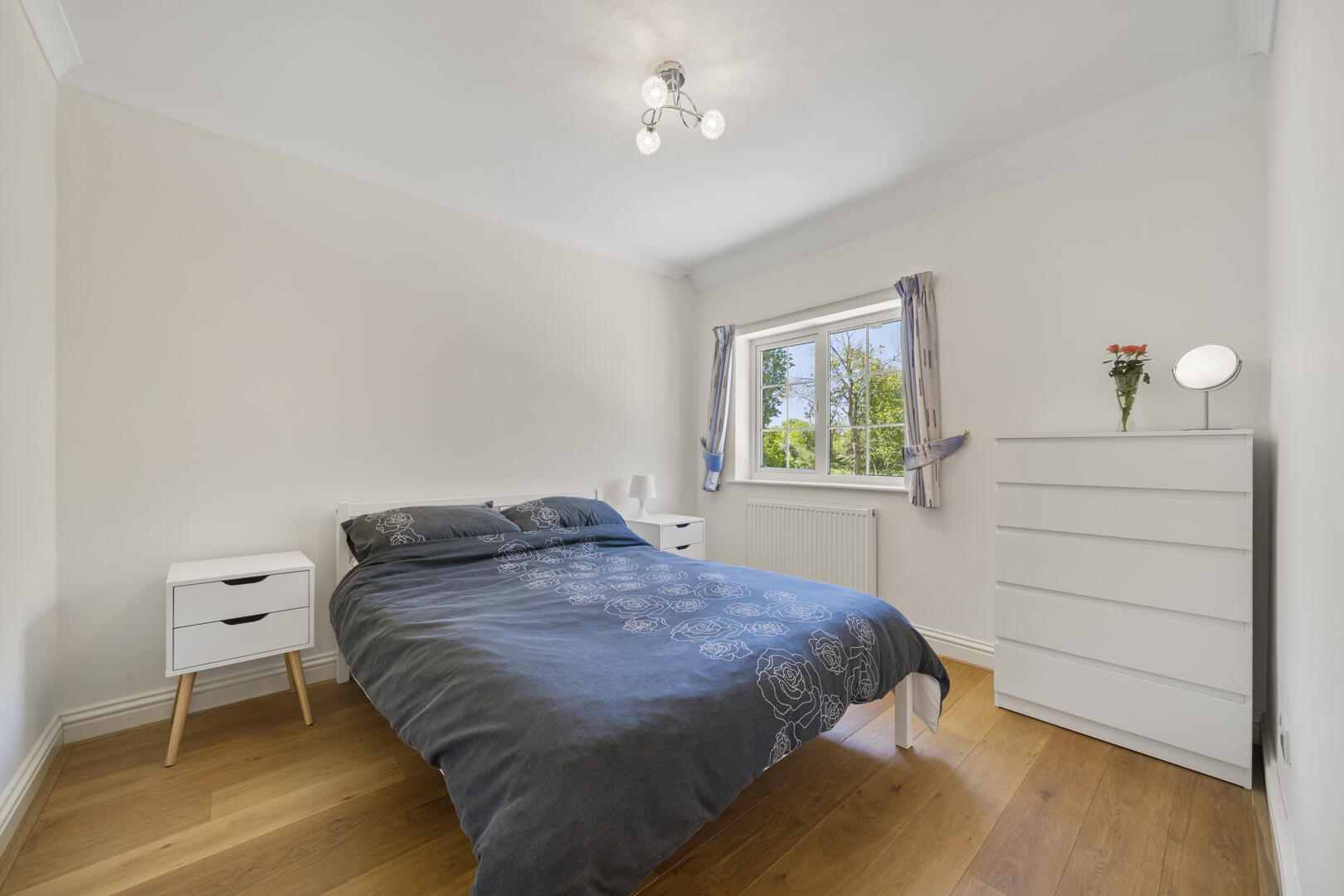 Bright minimalist bedroom with a double bed in blue floral bedding, two white nightstands, and a white dresser by the window.