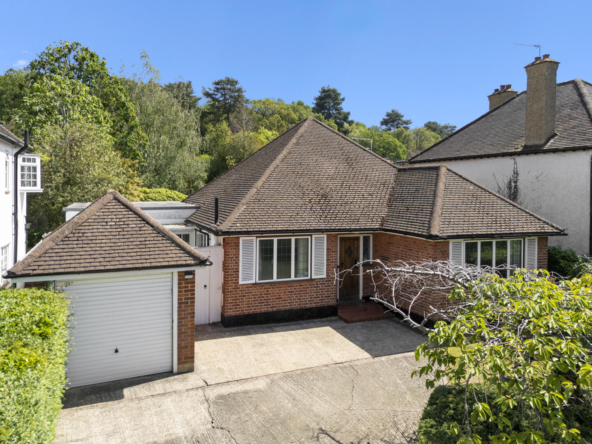 Front view of a single-story brick house with a brown tiled roof, white shutters, a attached garage, and a paved driveway surrounded by greenery.