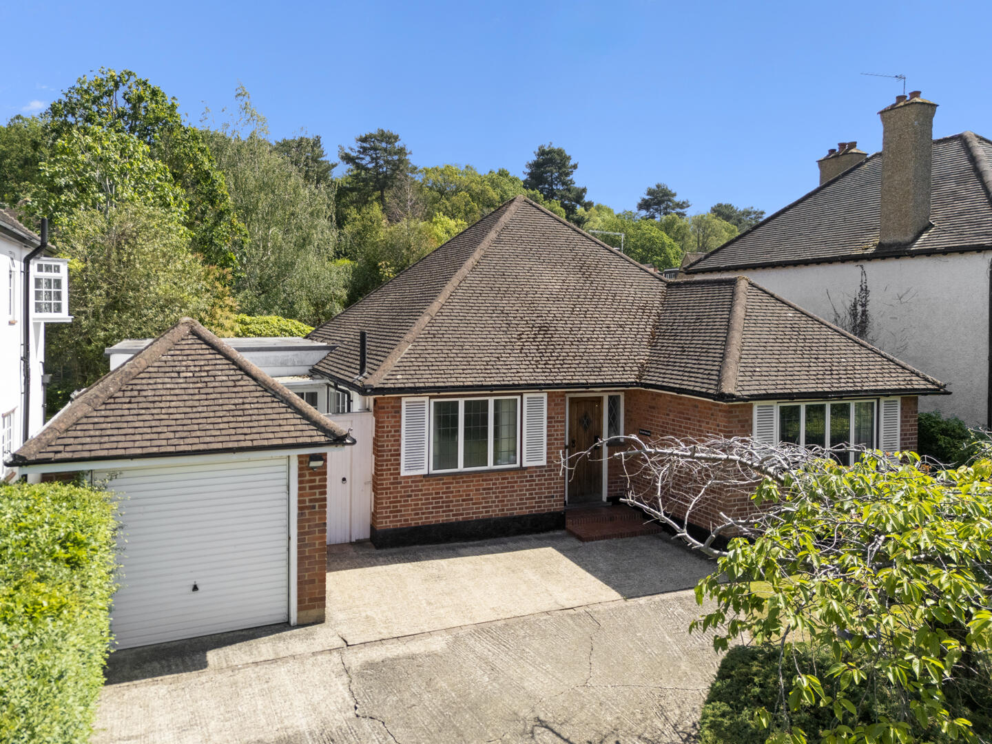 Front view of a single-story brick house with a brown tiled roof, white shutters, a attached garage, and a paved driveway surrounded by greenery.