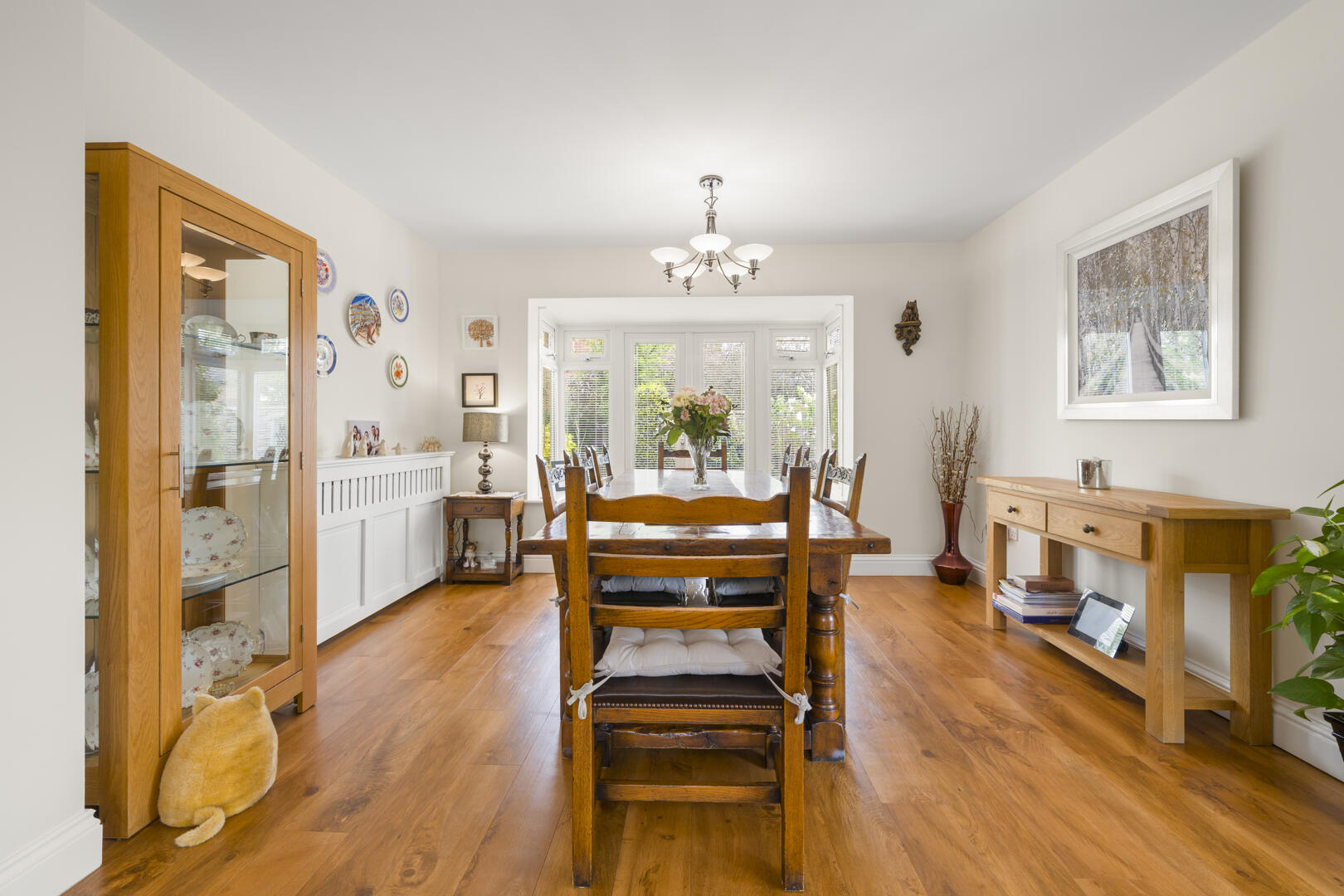 Wood dining room with a long wooden table, matching chairs, and a bouquet centerpiece by a bay window; warm wood floors and light fixture overhead.