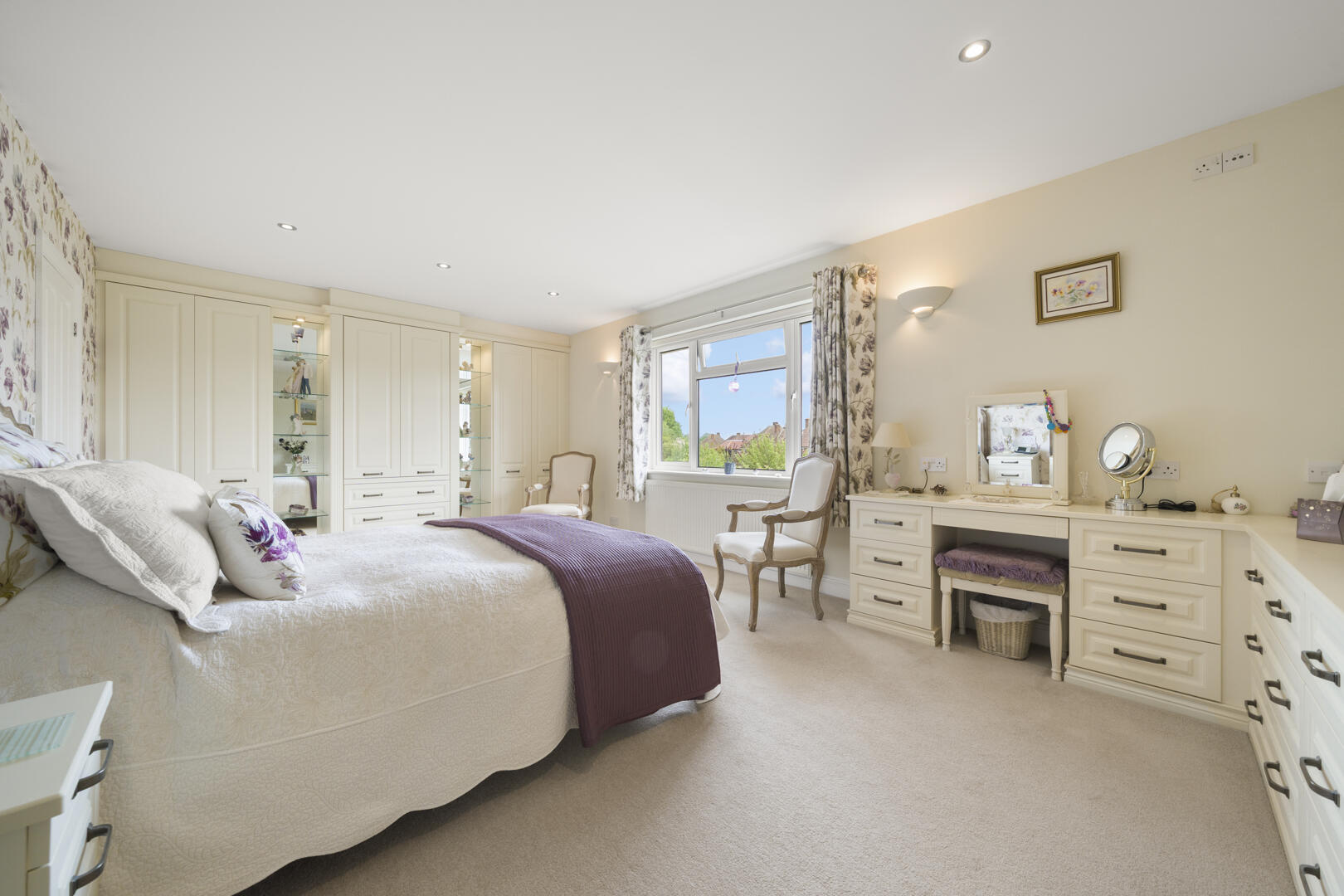 Bright cream-bedroom featuring built-in wardrobes, a large bed with white bedding and a purple throw, and a window with floral curtains across from a vanity desk.