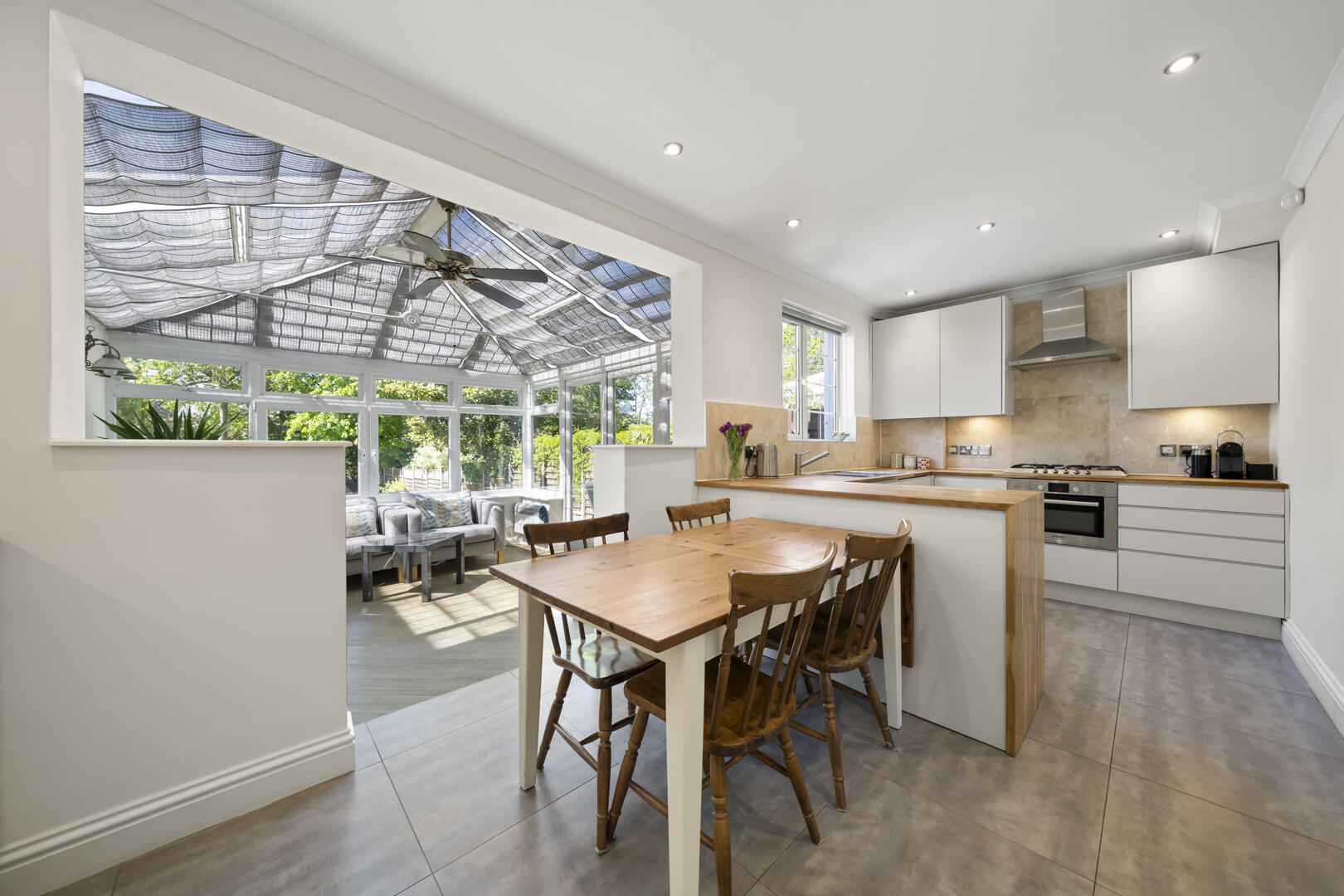 Open-plan kitchen with a wooden dining table and four chairs, leading to a bright conservatory with a ceiling fan.