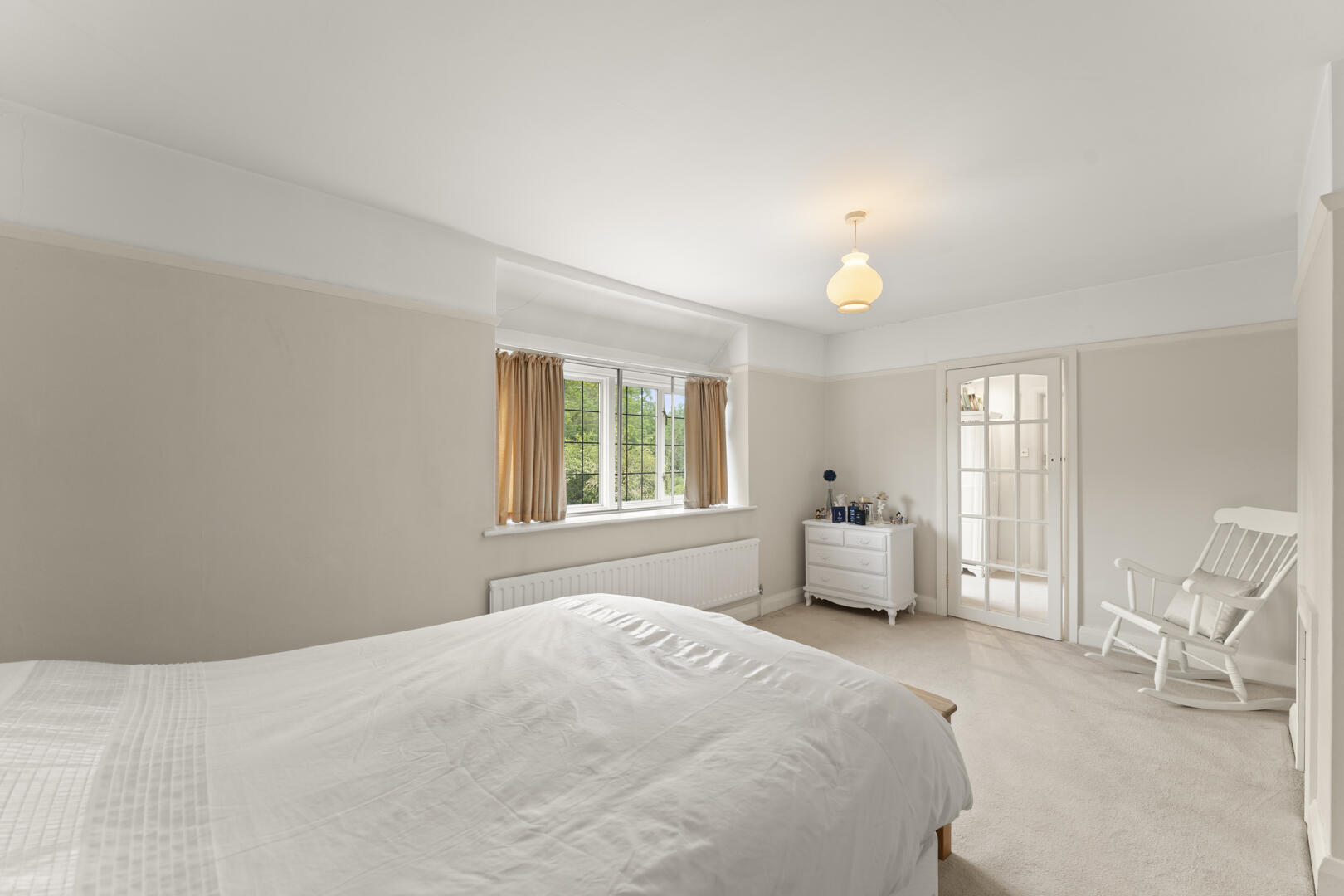 Bright bedroom with white bedding, beige curtains, and a large window overlooking greenery; white dresser, radiator, and a white rocking chair.