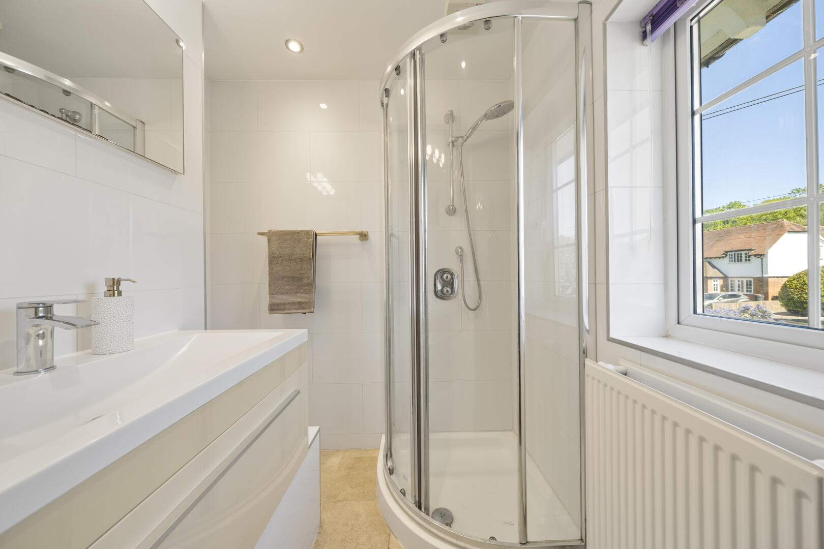 Bright white bathroom with a curved glass shower enclosure, white vanity, and a large window showing houses outside.