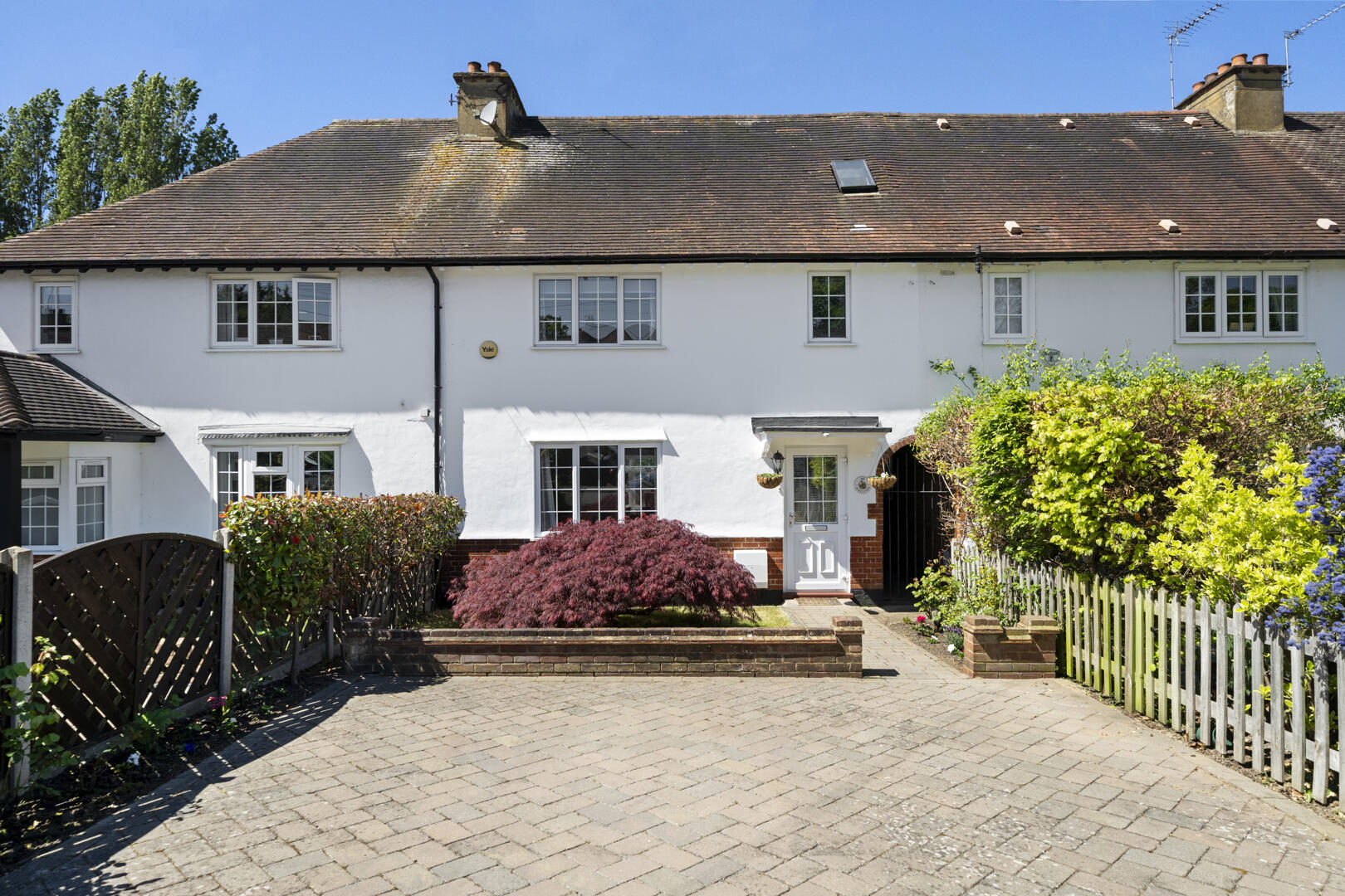 White two-story house with a red-tiled roof, front garden with a red shrub, and a paved driveway under a blue sky