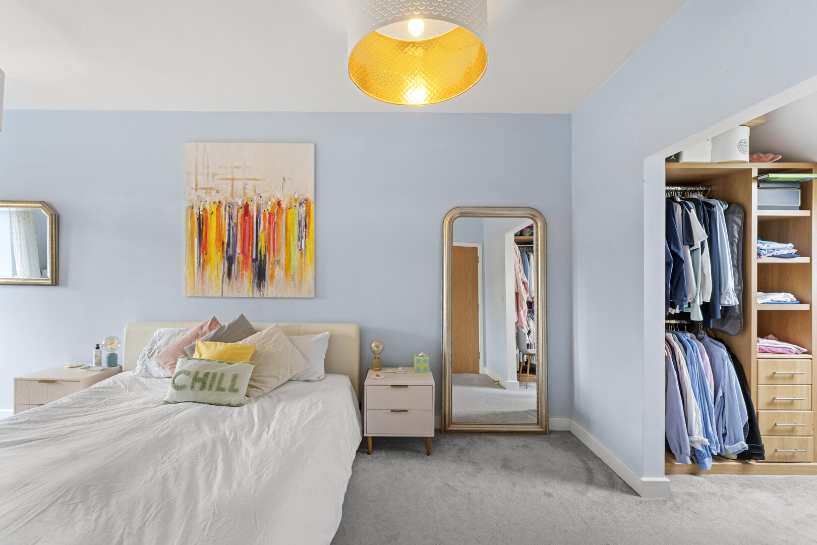 Bright bedroom with a white bed, pastel pillows, abstract wall art, and a tall mirrored wardrobe on a light blue wall.