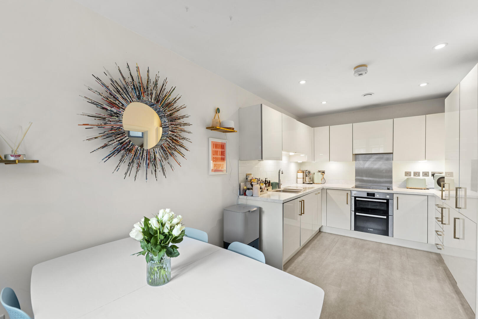 Modern white kitchen with glossy cabinets, stainless oven, and light gray floor; round sunburst mirror on the wall above a dining table with flowers in a glass vase.