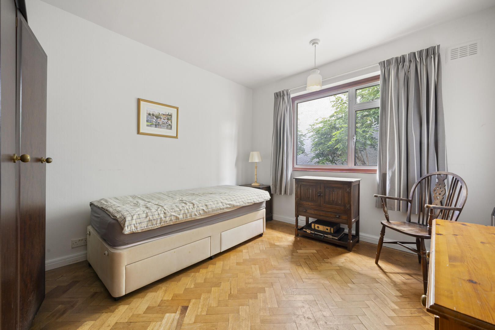 Small bedroom with a single bed along the left wall, plaid duvet, wooden wardrobe, and a window with gray curtains.