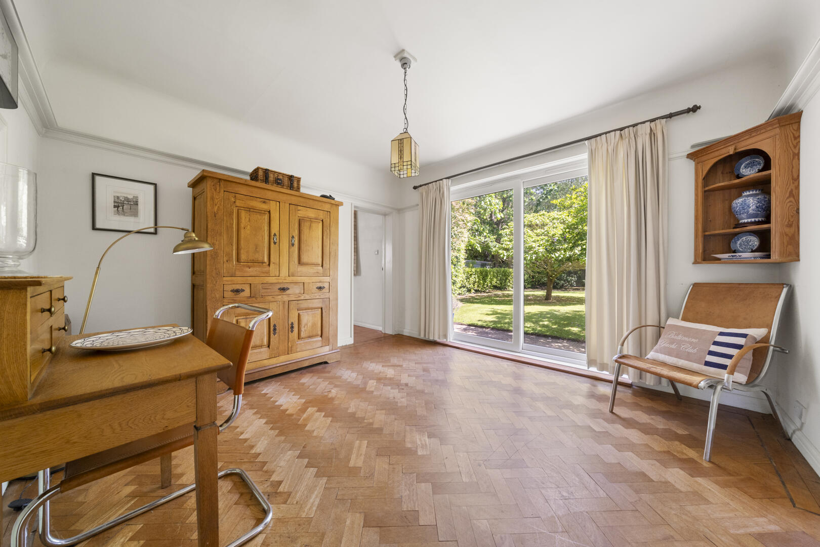 Sunlit room with parquet floor, a large wooden cabinet, desk, and a mid-century chair, opening to a green garden through sliding glass doors.