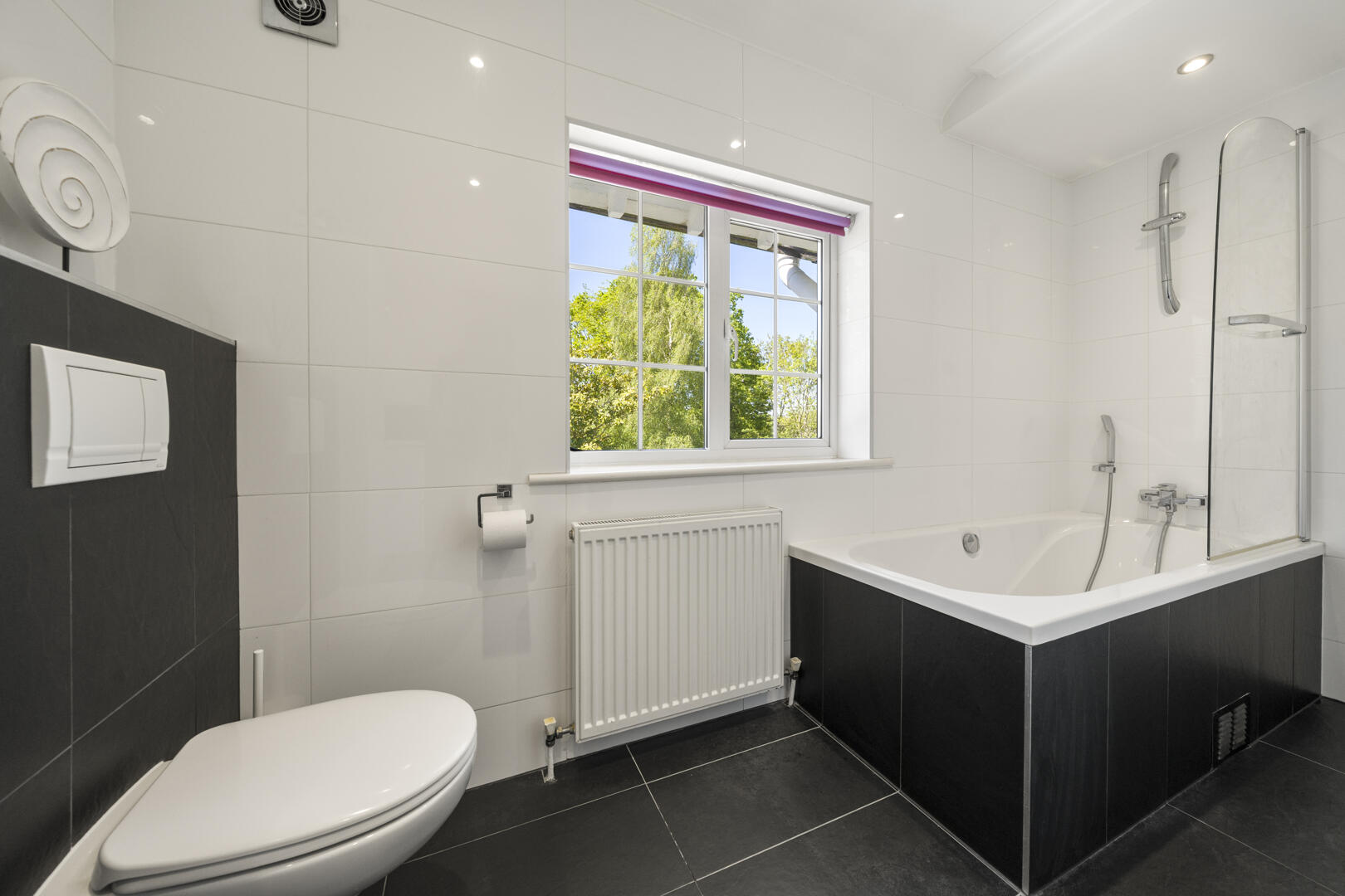 Modern bathroom with white tiles, a white bathtub with a black surround and glass screen, and a window view of trees outside.