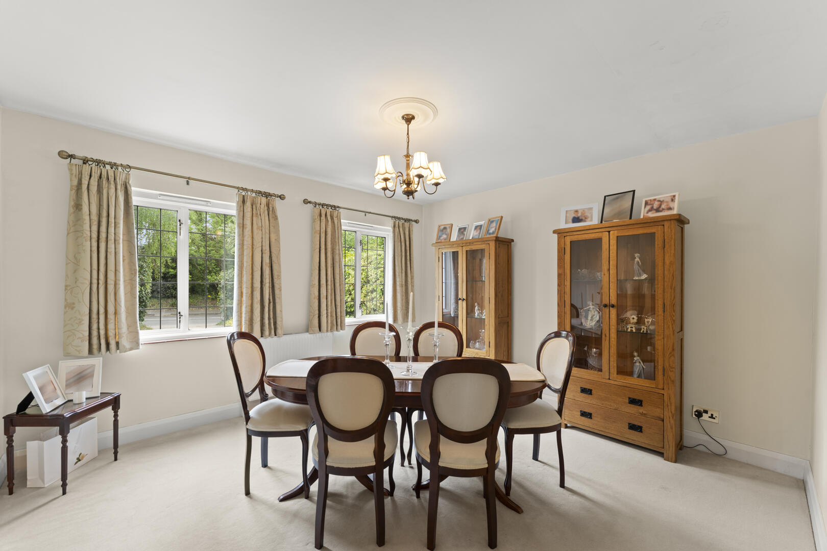 Traditional dining room with an oval wooden table and six upholstered chairs, beige walls, and a brass chandelier above. Two glass-front cabinets display china and photos, with large windows and patterned curtains in the background.
