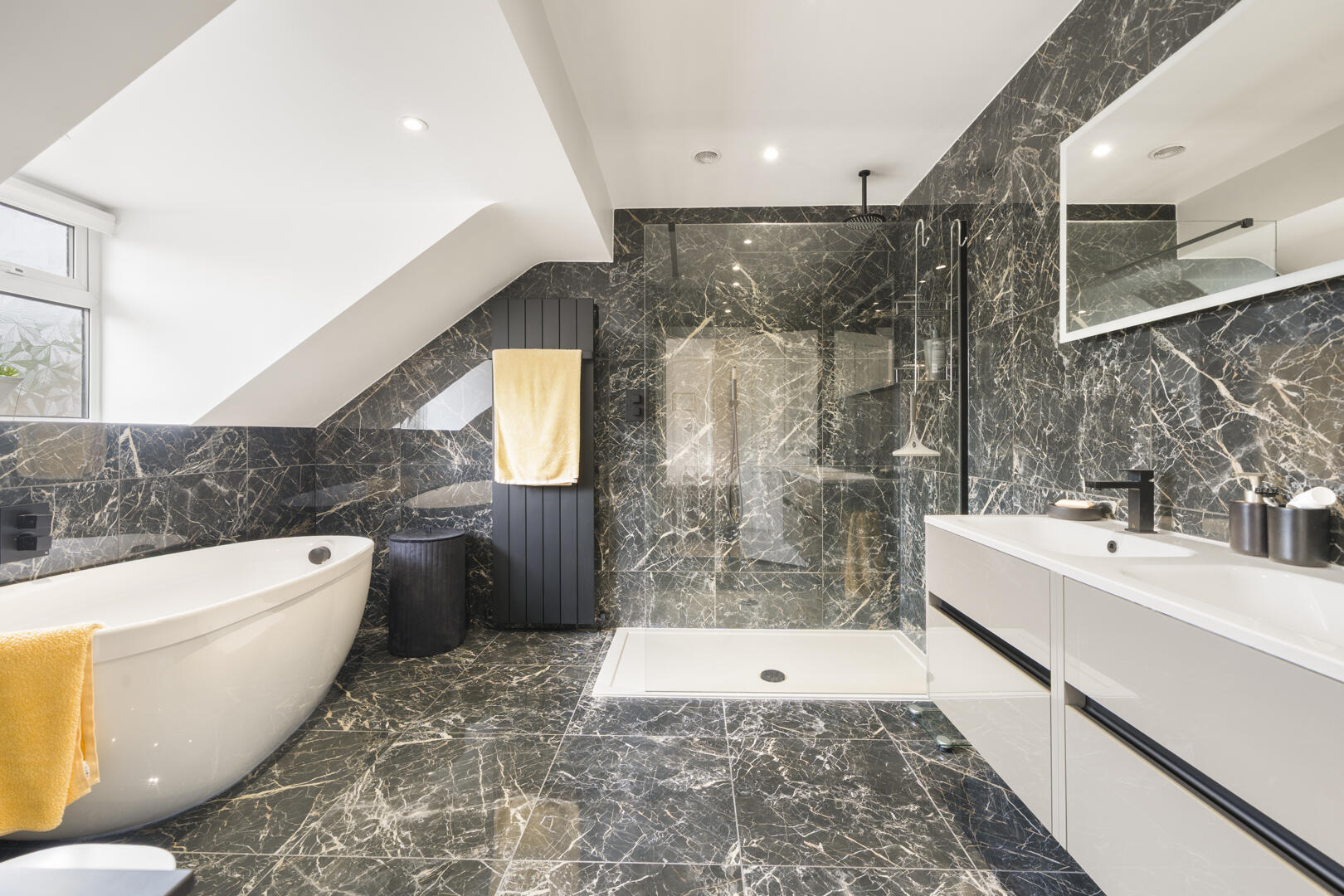 Luxurious bathroom with black marble walls and floor, a freestanding white tub, glass shower, and white double vanity with yellow towels nearby.