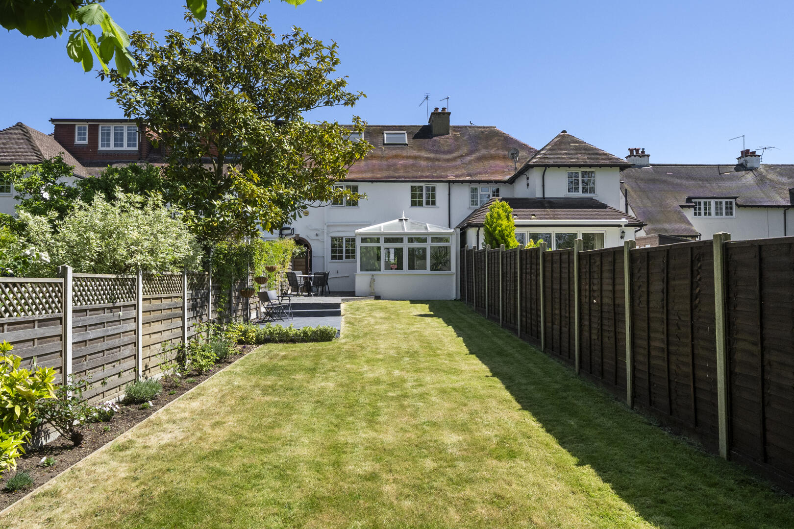 Backyard with a well-kept lawn, wooden fences on both sides, and a white house with a glass conservatory at the far end. Garden path and seating area visible on the left.