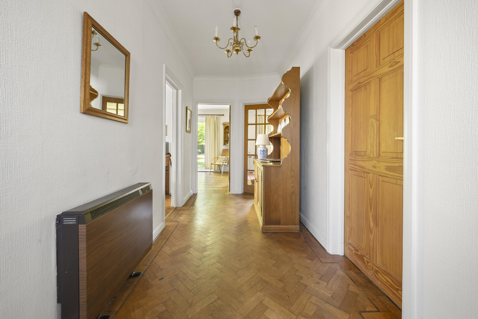 Bright hallway with herringbone wooden floor, white walls, and a brass chandelier overhead; doorway to rooms on both sides, and a wooden cabinet with a blue-and-white lamp to the right