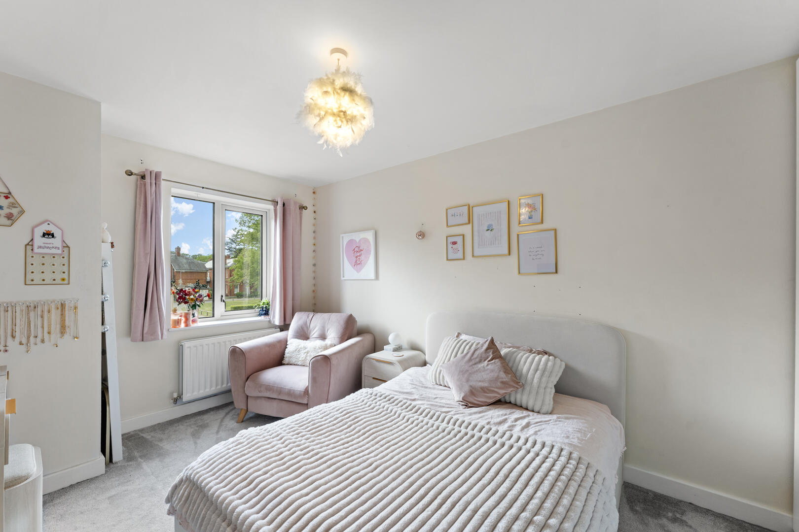 Soft pink bedroom with a plush bed, pink cushions, and a cozy pink chair by a window with pink curtains. Grounded in natural light and neutral decor.