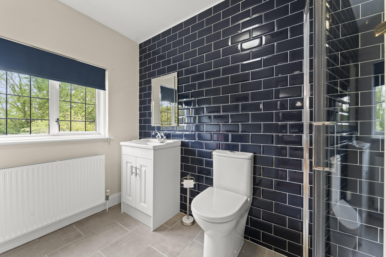 Modern bathroom with dark navy subway-tile wall, white vanity, toilet, and glass shower enclosure under a large window with blue blind