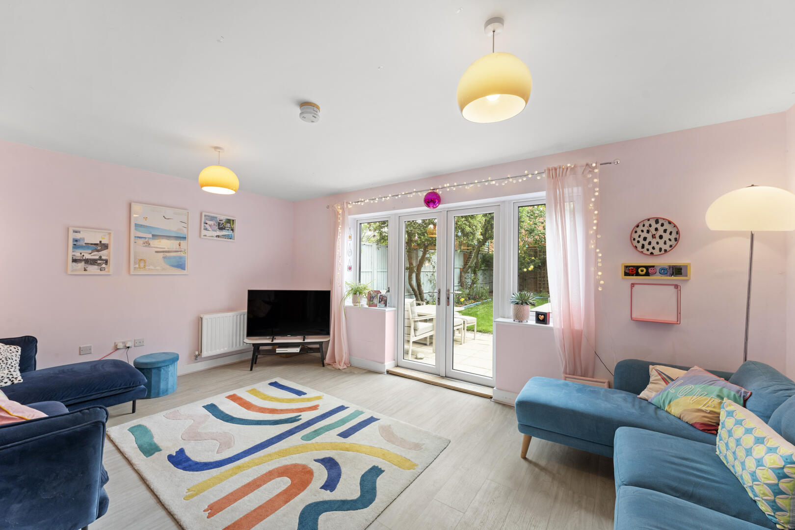 Pastel pink living room with blue sofas, a colorful abstract rug, and glass doors opening to a green garden with string lights above the window.