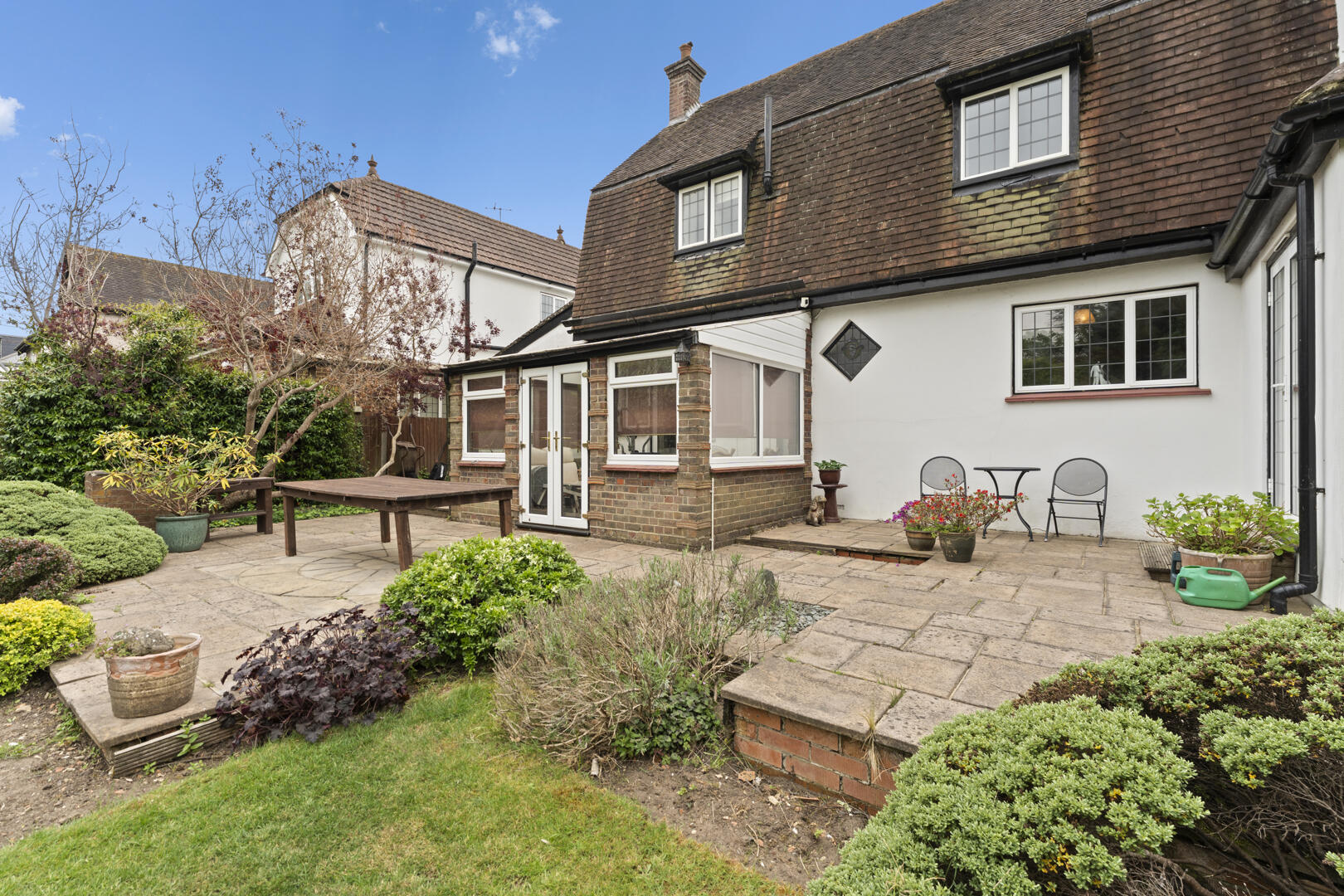Rear garden of a brick house with a paved patio, wooden table, two metal chairs, and potted plants.