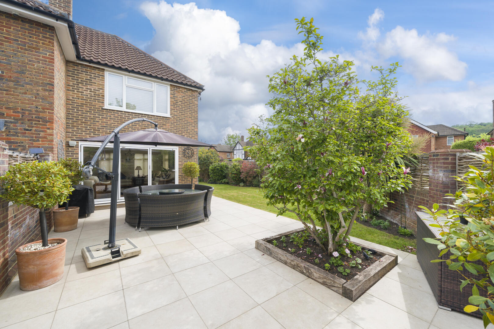 Backyard patio beside a brick house with sliding doors, a round wicker seating set under a large umbrella, and a raised planter with a tree.
