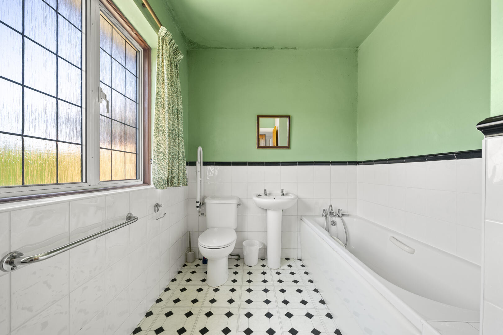 Bright bathroom with green walls above white tile wainscoting, a white toilet, pedestal sink, and a built-in bathtub along a black-and-white checkered floor.