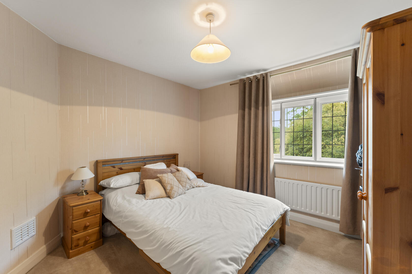 Cozy bedroom with a wooden bed, bedside table and lamp, beige patterned walls, and a large window with brown curtains