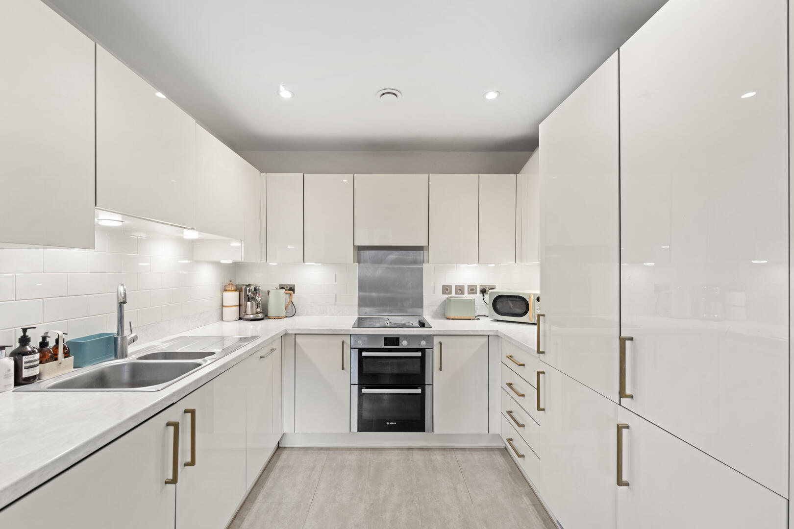 Modern white U-shaped kitchen with glossy cabinets, marble countertops, and a stainless steel oven.