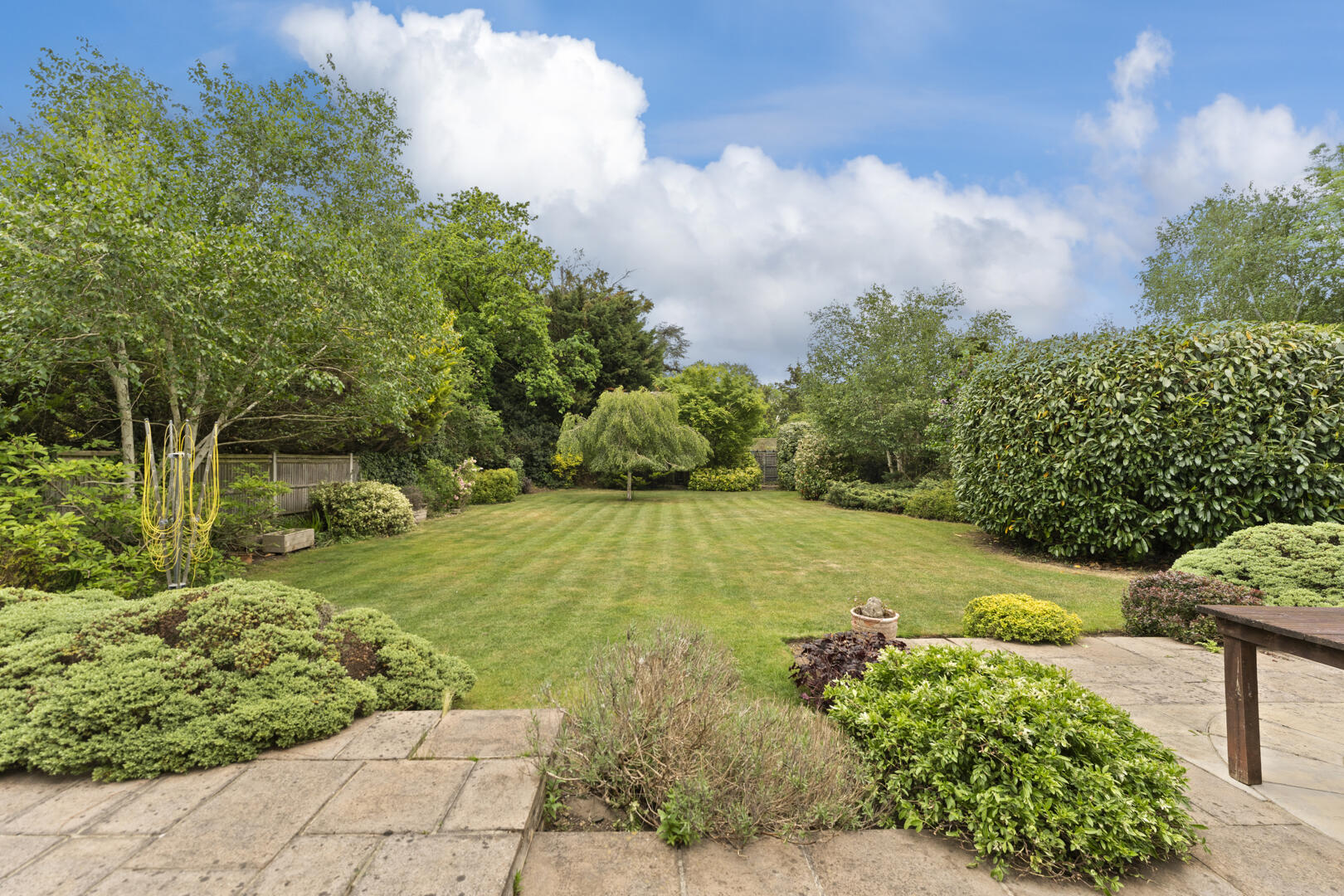 Formal backyard garden with a neatly mowed lawn, stone patio in the foreground, and trimmed shrubs along a wooden fence.